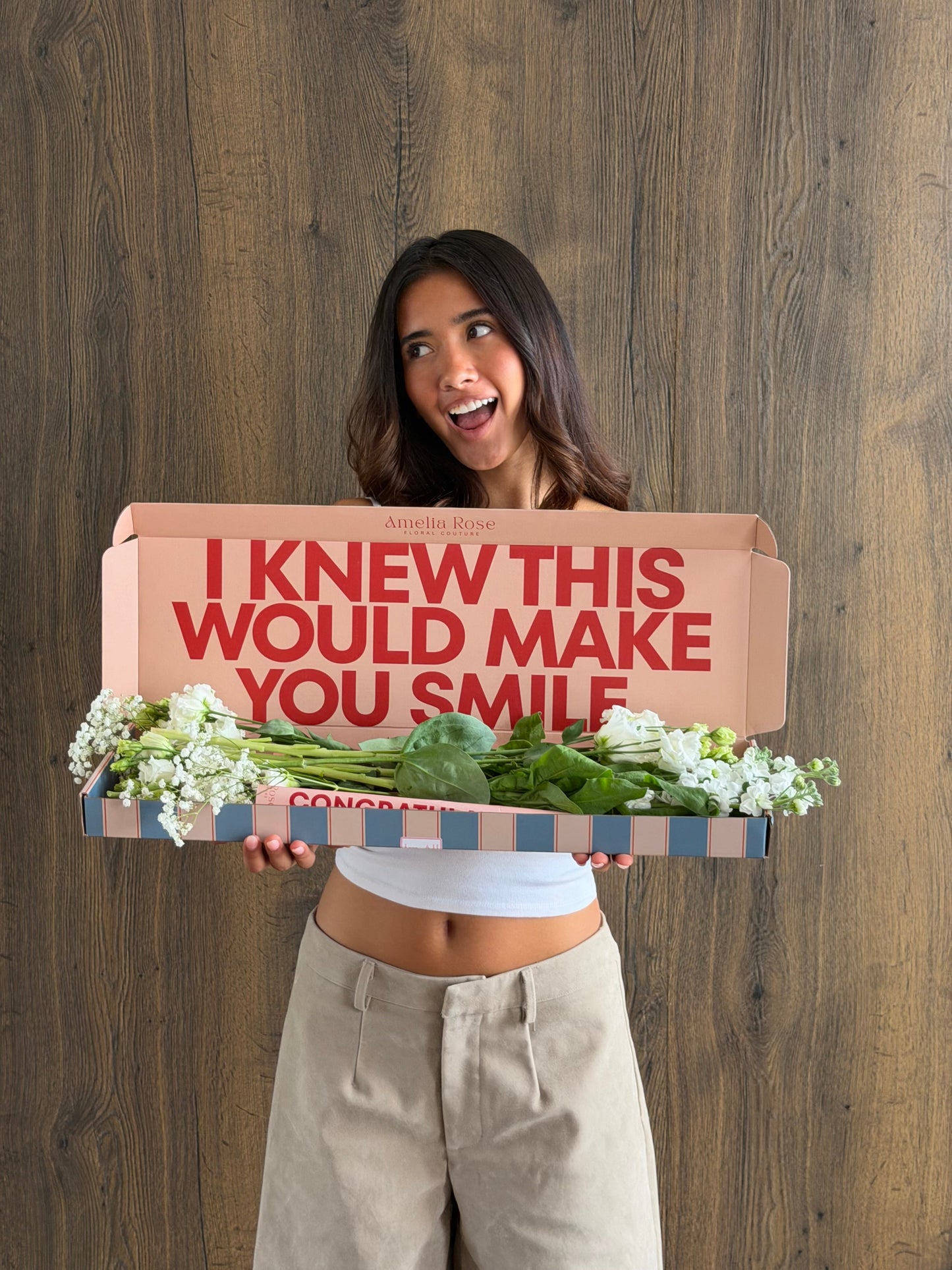 A woman holding a surprise gift box with the text 'I KNEW THIS WOULD MAKE YOU SMILE' in red. The box contains a striped bag of 'Congratulations' branded produce and white flowers.